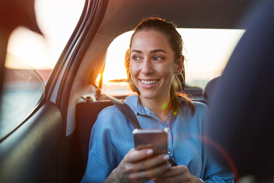 About Our Agency - Young Woman Uses a Cell Phone in the Back Seat of a Car, Smiling