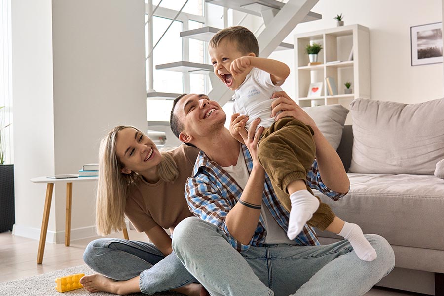 Personal Insurance - Mother and Father Sit Cross-Legged on Their Living Room Floor, Lifting Their Toddler Son Overhead, All Smiling